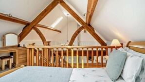 Een slaapkamer met schuine wanden, houten balken, een bed met bloemenbeddels, een bureau met spiegel, en een nachtkastje met lamp.