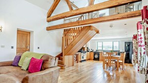 Een ruime woonkamer met een houten trap, een bank met kleurrijke kussens, een eettafel en een keuken op de achtergrond.