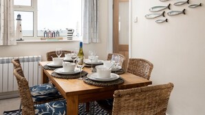 Uma sala de jantar com uma mesa de madeira posta para quatro pessoas, rodeada por cadeiras de vime, e vista para o mar através de uma janela.