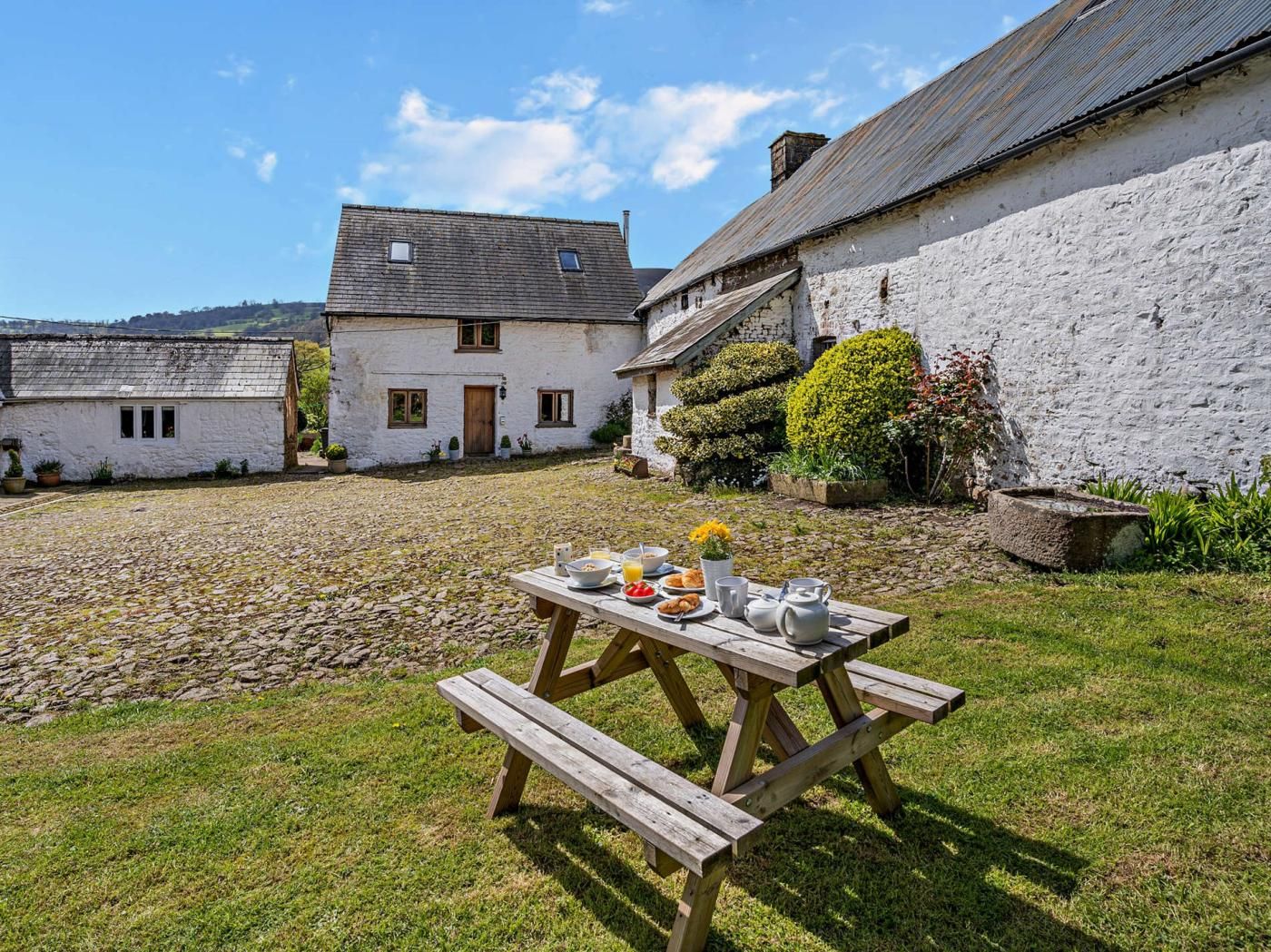 A courtyard with a picnic table set up with food and drinks, surrounded by white buildings with stone walls.