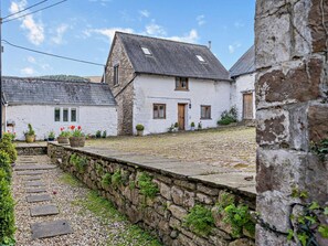A stone wall leading to a white building with a sloped roof, a wooden door, and a window with a shutter.