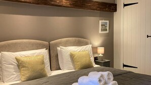 Cama doble con colcha gris, dos almohadas blancas y una almohada decorativa dorada. Hay dos toallas enrolladas sobre la cama.