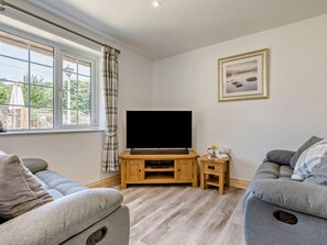 A living room with a flat-screen TV on a wooden stand, two grey sofas, a small side table with a book and a cup, and a window with curtains. - 2 Bed in Looe (oc-90966) (Trelawne)