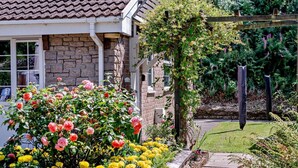Uma casa com fachada de pedra, telhado de telhas e um jardim com flores e uma pérgula de madeira.