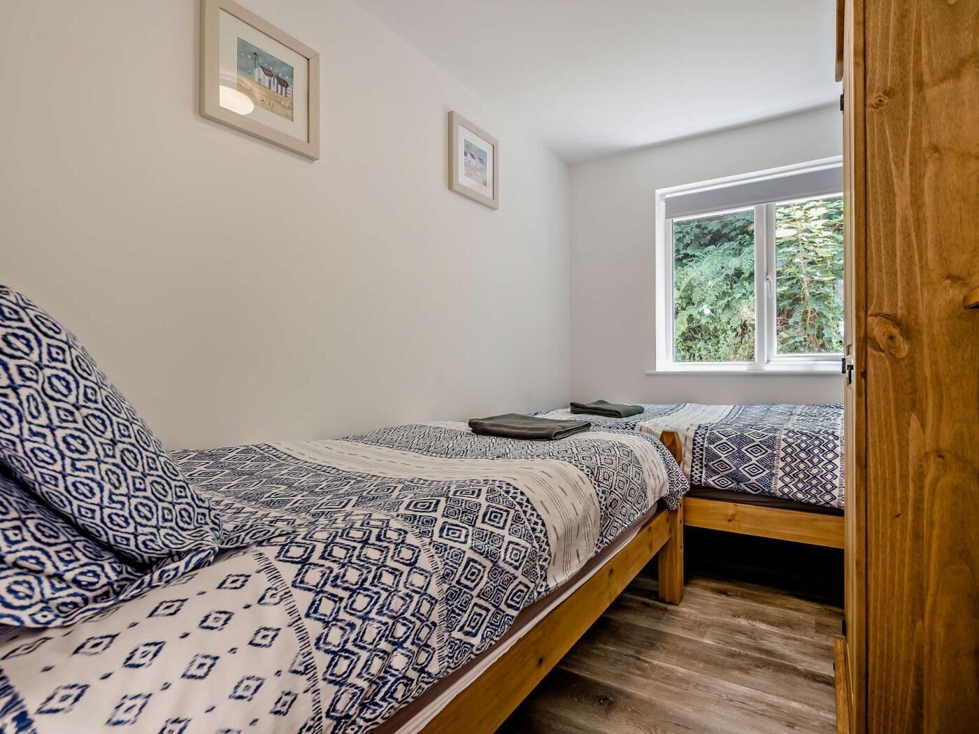 A bedroom with two beds, a window with a view of greenery, and framed pictures on the wall.