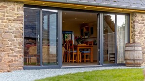 A stone building with a glass door leading to a dining area with wooden furniture and a table. - 3 Bed in Ratlinghope (oc-79238) (Ratlinghope)
