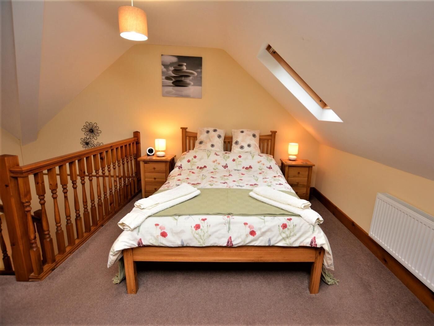 Een slaapkamer met een schuine plafond, een houten bedframe, een bloemenbedekking, twee bedlampen en een kleine wanddecoratie.
