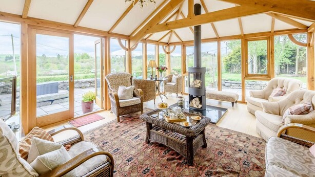 A spacious room with a high ceiling, large windows, wicker furniture, and a central fireplace. - 6 Bed in Bala (oc-80348) (Fron-goch)