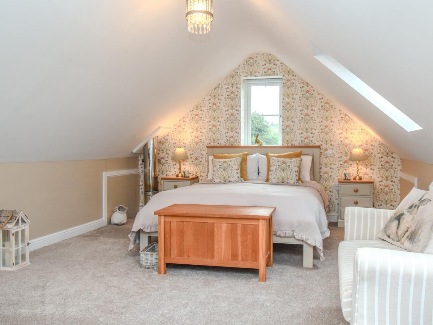 A bedroom with a sloped ceiling, a bed with matching headboard, bedside tables with lamps, a wooden bench, and a window with a view.
