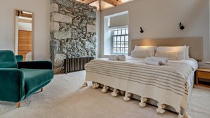Ein Schlafzimmer mit Steinwand, einem Bett mit gestreifter Bettwäsche, einem grünen Sessel und einer Holzbalkendecke.