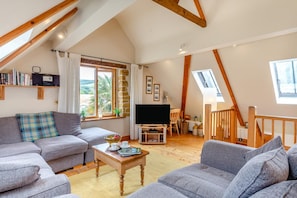 A cozy living room with a grey sofa, a coffee table, and a TV. There are wooden beams on the ceiling and a window with a view of the outdoors. - 2 Bed in Bridport (oc-dc111) (Chideock)