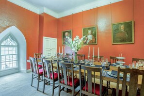 A dining room with a large table set for a meal, surrounded by chairs with red cushions, and framed portraits on the walls.