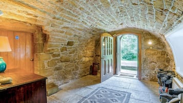 A stone-walled corridor with a wooden door leading outside, a lamp, and a rug on the floor.