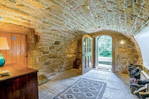 A stone-walled corridor with a wooden door leading outside, a lamp, and a rug on the floor.