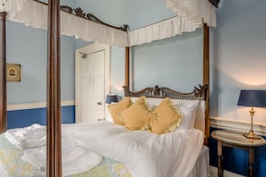 A four-poster bed with white linens and two beige pillows, a bedside table with a lamp, and a framed picture on the wall.