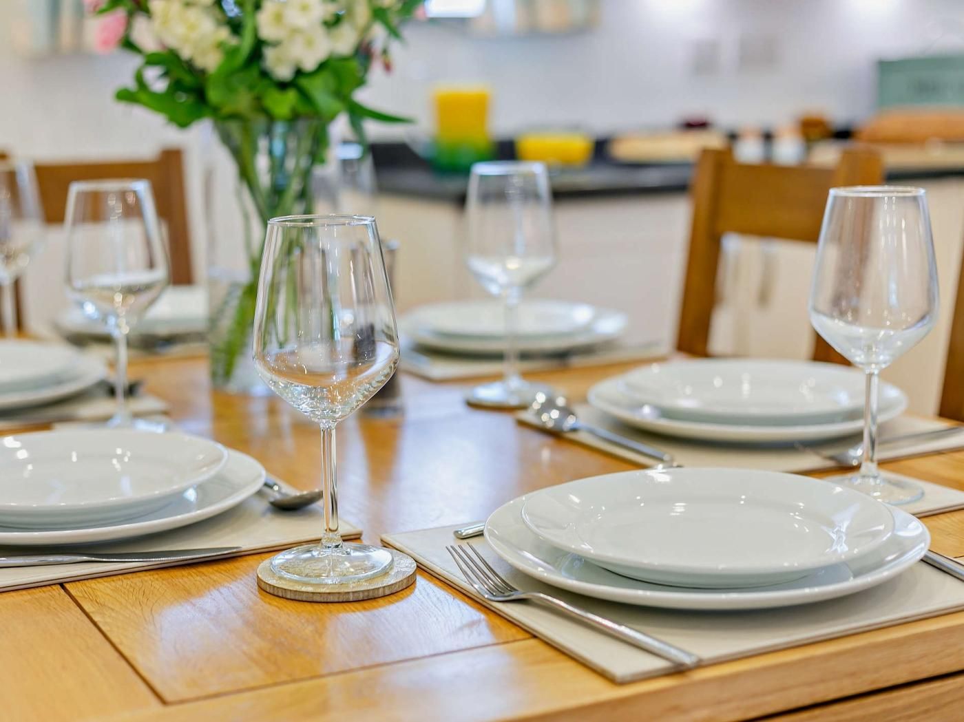 A dining table set with plates, cutlery, and wine glasses, with a vase of flowers in the background.