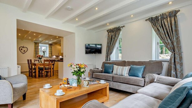 A living room with a sofa, a coffee table, and a TV. There is a dining area with a table and chairs visible in the background. - 3 Bed in Hayle (oc-fodry) (Saint Erth)