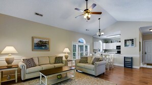 A living room with a sofa, coffee table, and lamps. There is a ceiling fan, a painting, and a kitchen in the background. - Unwind At The Riverwalk, Spacious 2BR Retreat (Myrtle Beach)