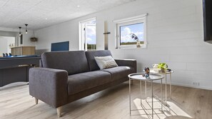 A modern living room with a grey sofa, a small round table, and a view of the outdoors through a window. - VillaVilla 967 - Bønnerup Strand, Østjylland (Glesborg)