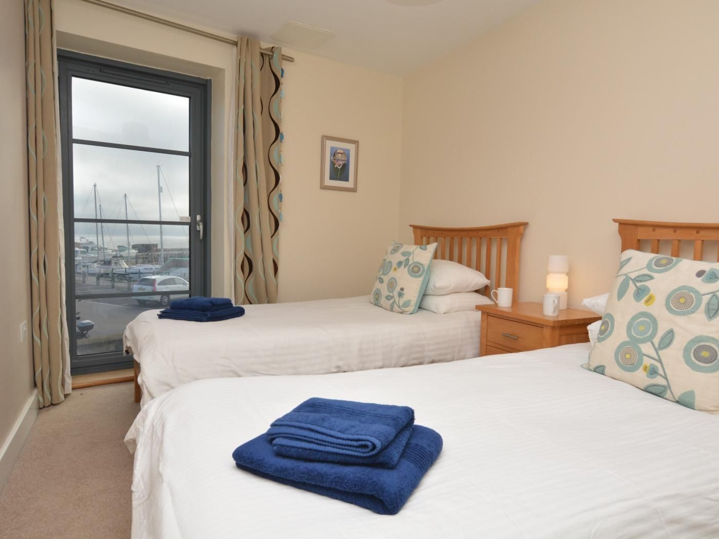 A hotel room with two beds, a window with a view of boats, a small bedside table with a lamp, and a wall-mounted picture.