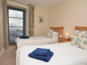 A hotel room with two beds, a window with a view of boats, a small bedside table with a lamp, and a wall-mounted picture. - 2 Bed in Bridport (oc-pebba) (West Bay)