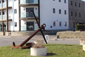 A concrete planter with a rusted anchor on a grassy area in front of a modern building with multiple windows and balconies. - 2 Bed in Bridport (oc-pebba) (West Bay)