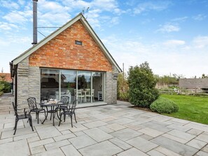 Et murstenshus med skorsten, en terrasse med udendørs møbler og en klar himmel.