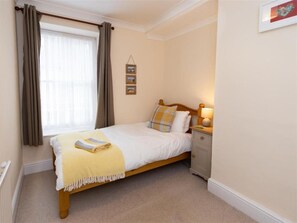 A neatly made bed with a yellow throw blanket, a bedside table with a lamp, and a window with curtains. - 3 Bed in Tenby (oc-fb141) (Tenby)