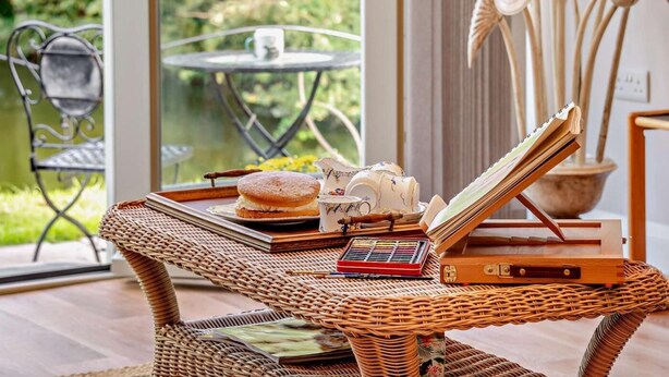 Une table en osier, avec un livre, une théière et un plateau sur lequel se trouve une pâtisserie.
