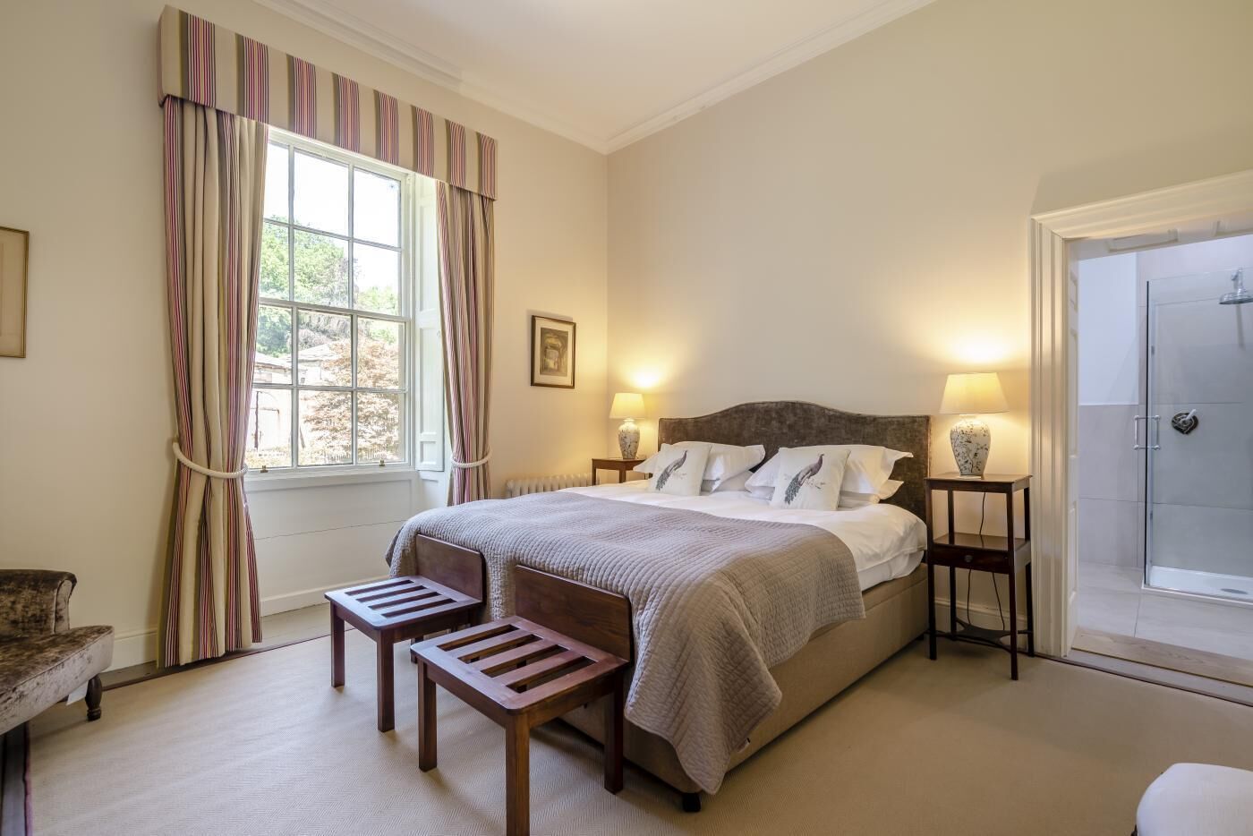 Een slaapkamer met een bed, nachtkastjes, een stoel, een raam met gordijnen, en een deur naar een douche.