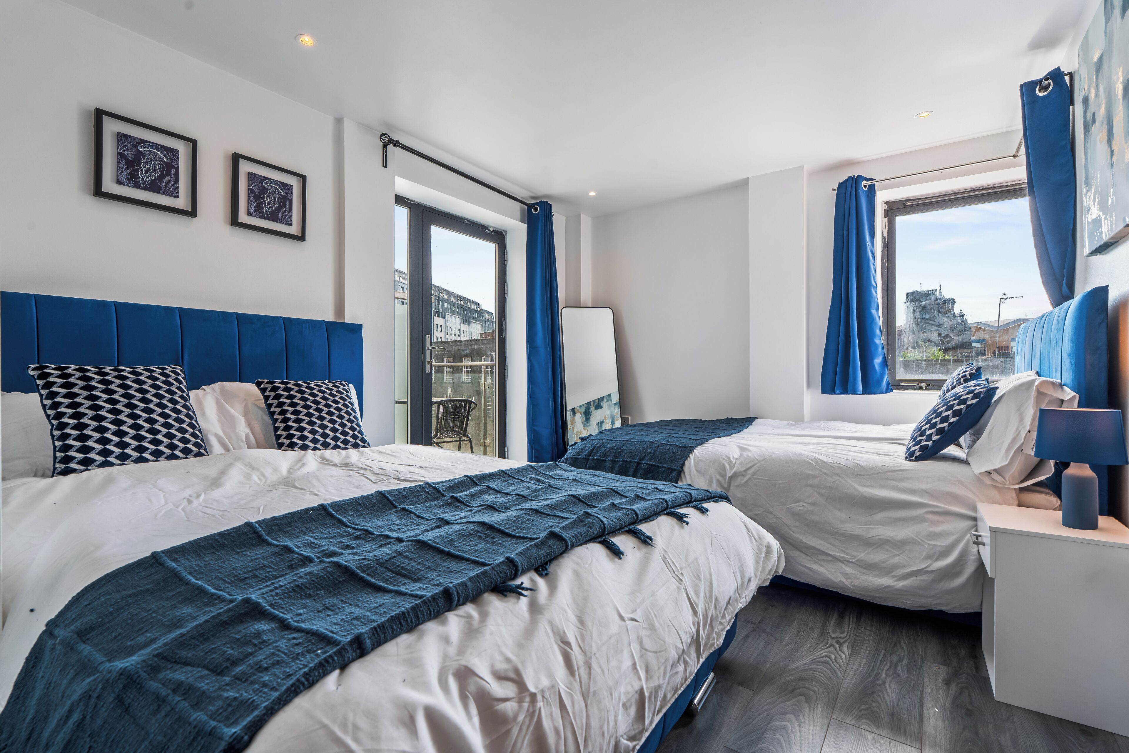 Une chambre d’hôtel moderne avec deux lits, une grande fenêtre avec des rideaux, une vue sur les immeubles et une tête de lit bleue.