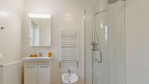 Un baño con ducha de vidrio, un bidé, un lavamanos blanco, un espejo y un perchero para toallas.