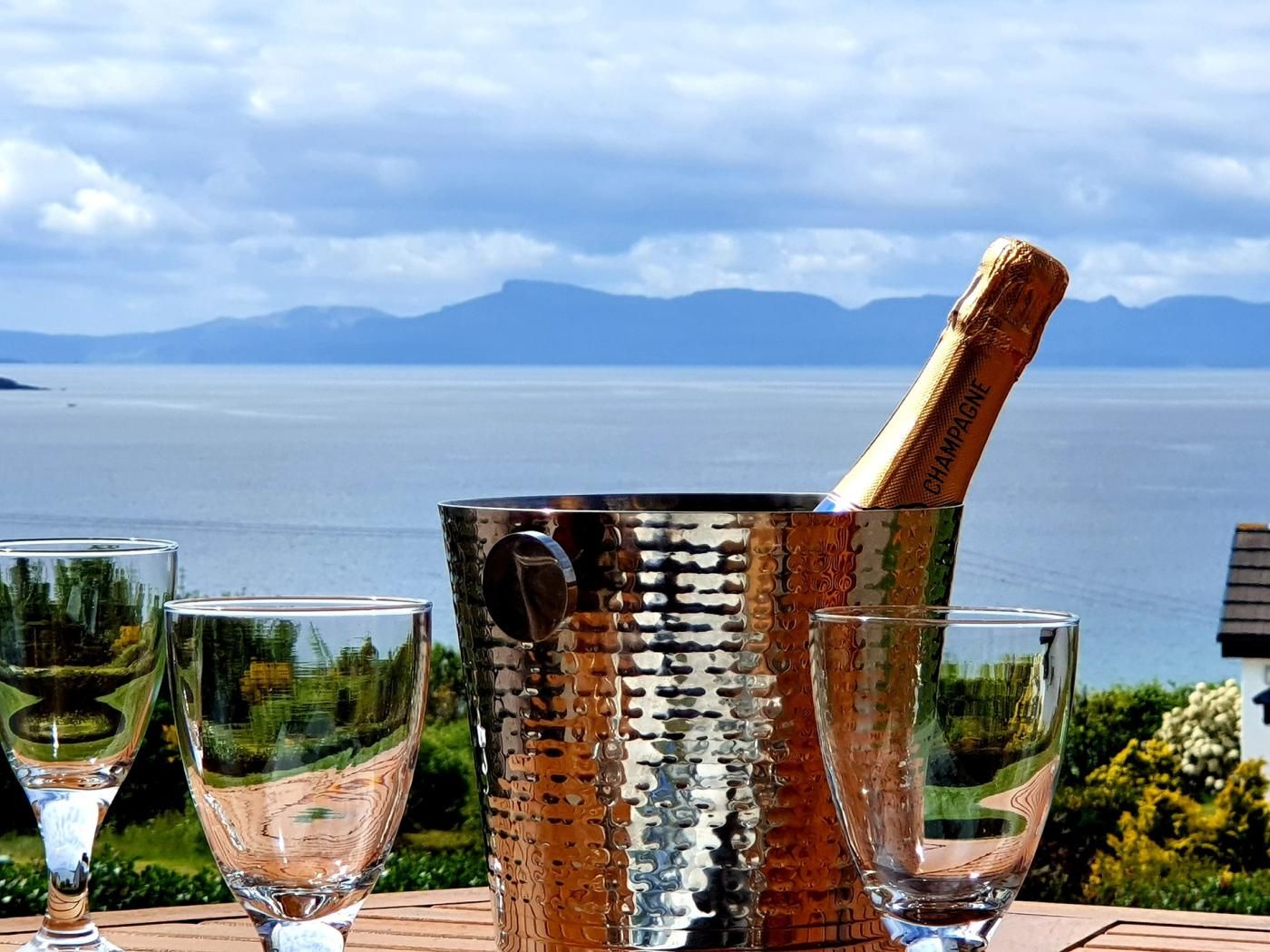 Un cubo metálico color champán con una botella sobre una mesa con tres copas, con vistas a un cuerpo de agua y montañas.