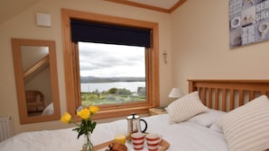 Un dormitorio con cama, ventana con vista al lago, mesita de noche con lámpara y bandeja con comida.