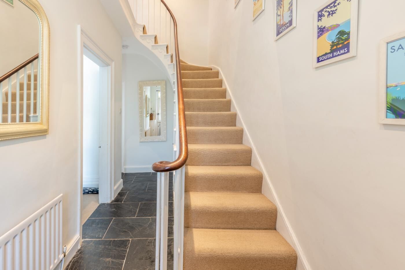 A staircase with a wooden handrail and beige carpeted steps, leading to a room with a white door and a mirror on the wall.
