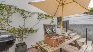 Mesa de picnic de madera con una cesta, una parrilla y un paraguas en un patio.