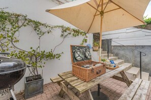 Ein Holztisch für ein Picknick mit Korb, Grill und Sonnenschirm auf einer Terrassenfläche.