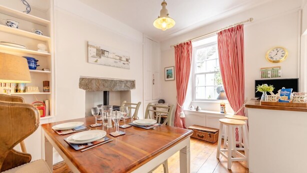 Un comedor con una mesa de madera dispuesta para cuatro, una zona de cocina con ventana, y un estante con utensilios de cocina.
