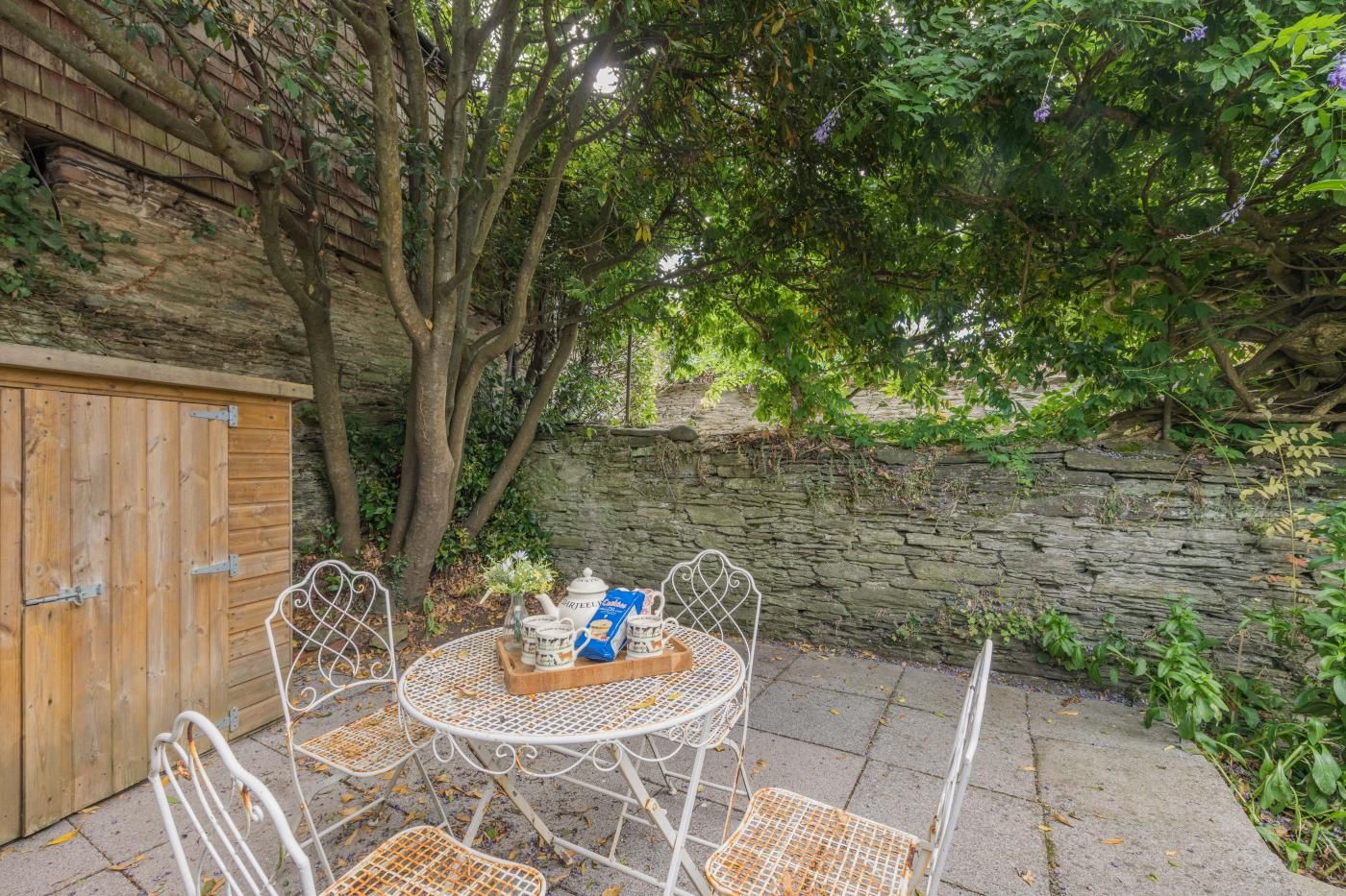 A patio with a round metal table and four chairs, a wooden gate, and a stone wall.
