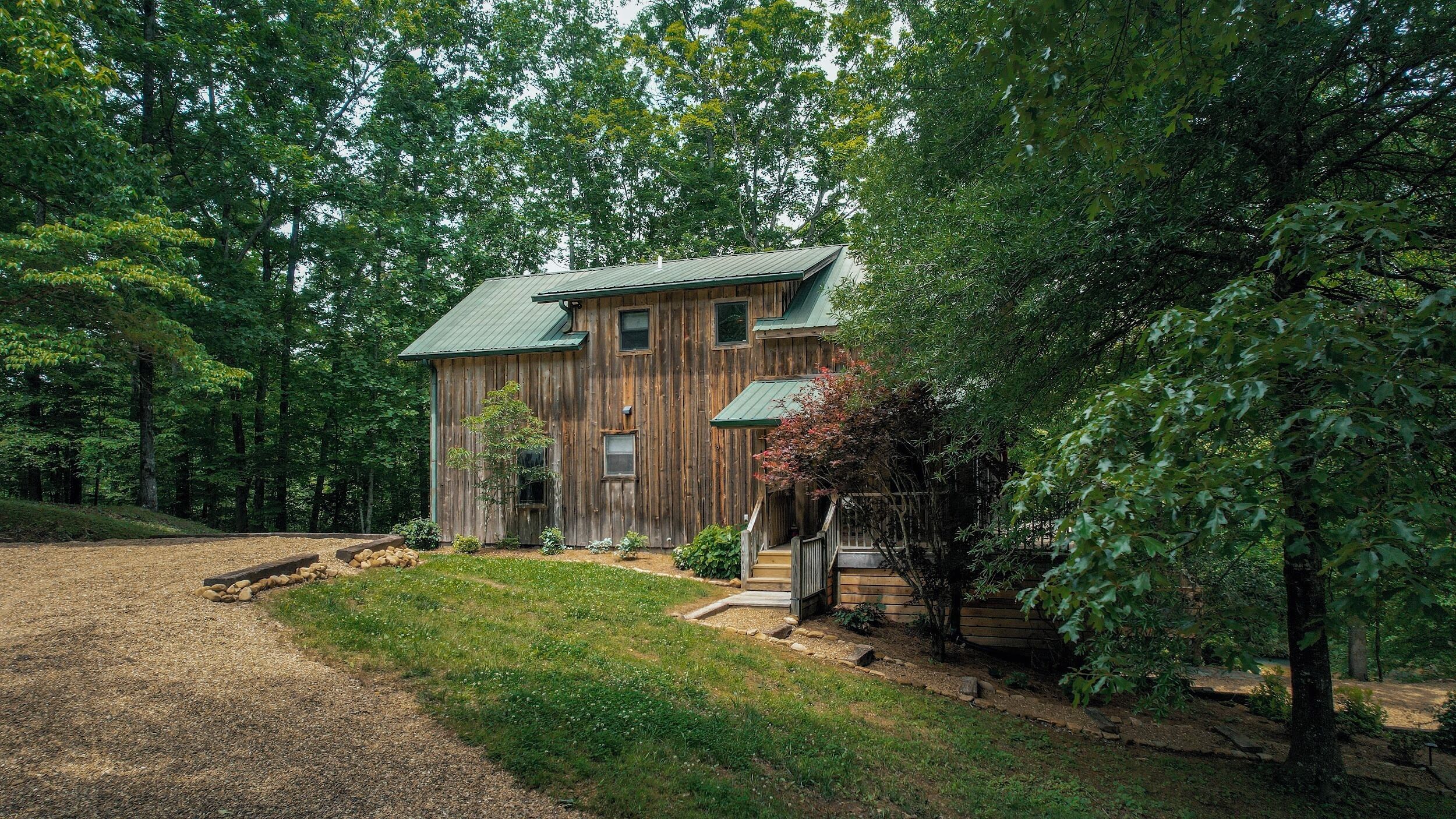 A rustic wooden house with a green roof, surrounded by dense trees and a gravel driveway.