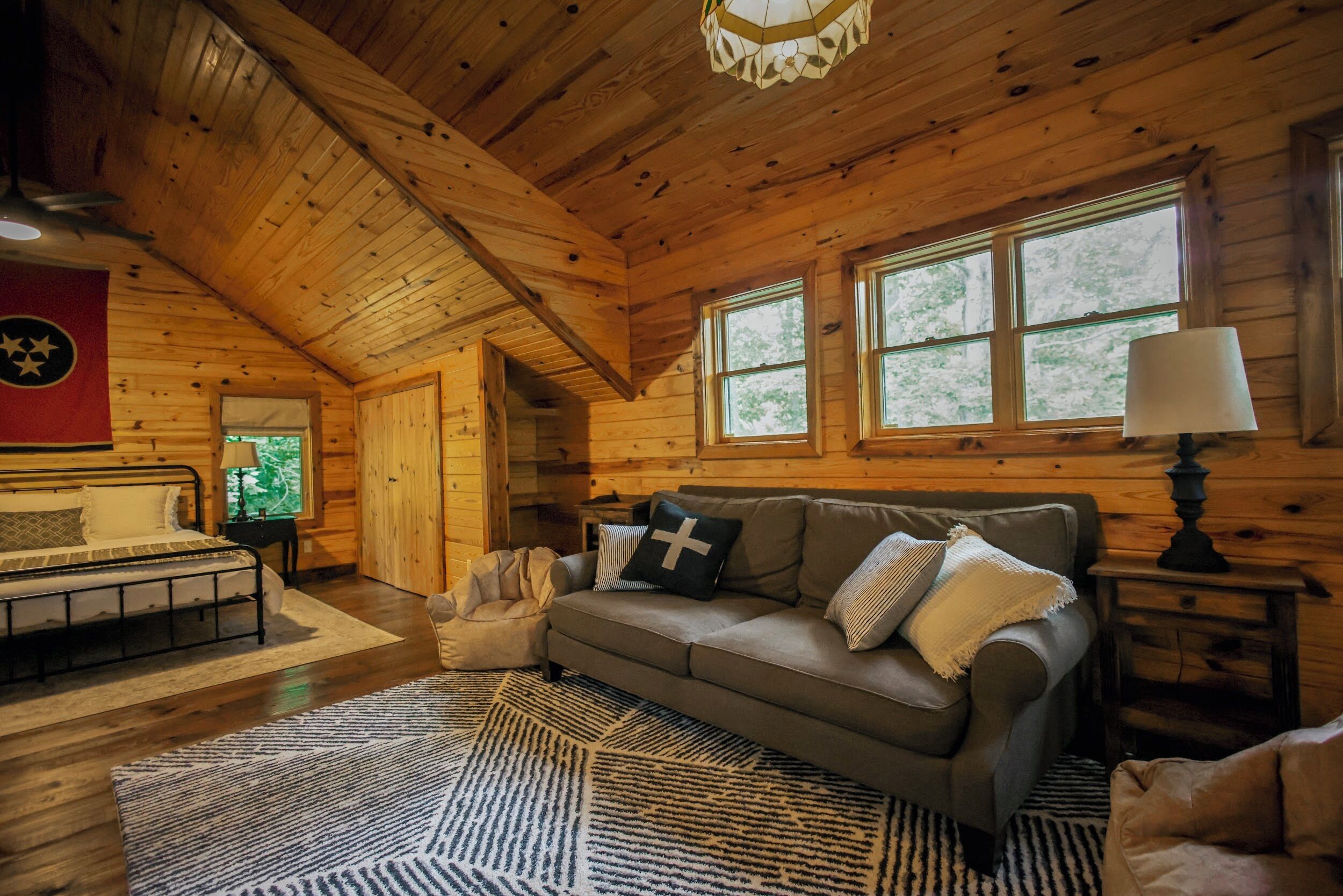 A cozy cabin interior with a wooden ceiling, a sofa with cushions, a patterned rug, and a window with a view of trees.