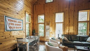 A cozy cabin interior with a dining area, a sofa, and a large window. - Private Cabin · Creekside · Hot tub · Fire pit · Only 1hr to Nashville (Nunnelly)