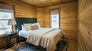 A wooden cabin bedroom with a bed, nightstand, lamp, and a window with a view of trees. - Private Cabin · Creekside · Hot tub · Fire pit · Only 1hr to Nashville (Nunnelly)