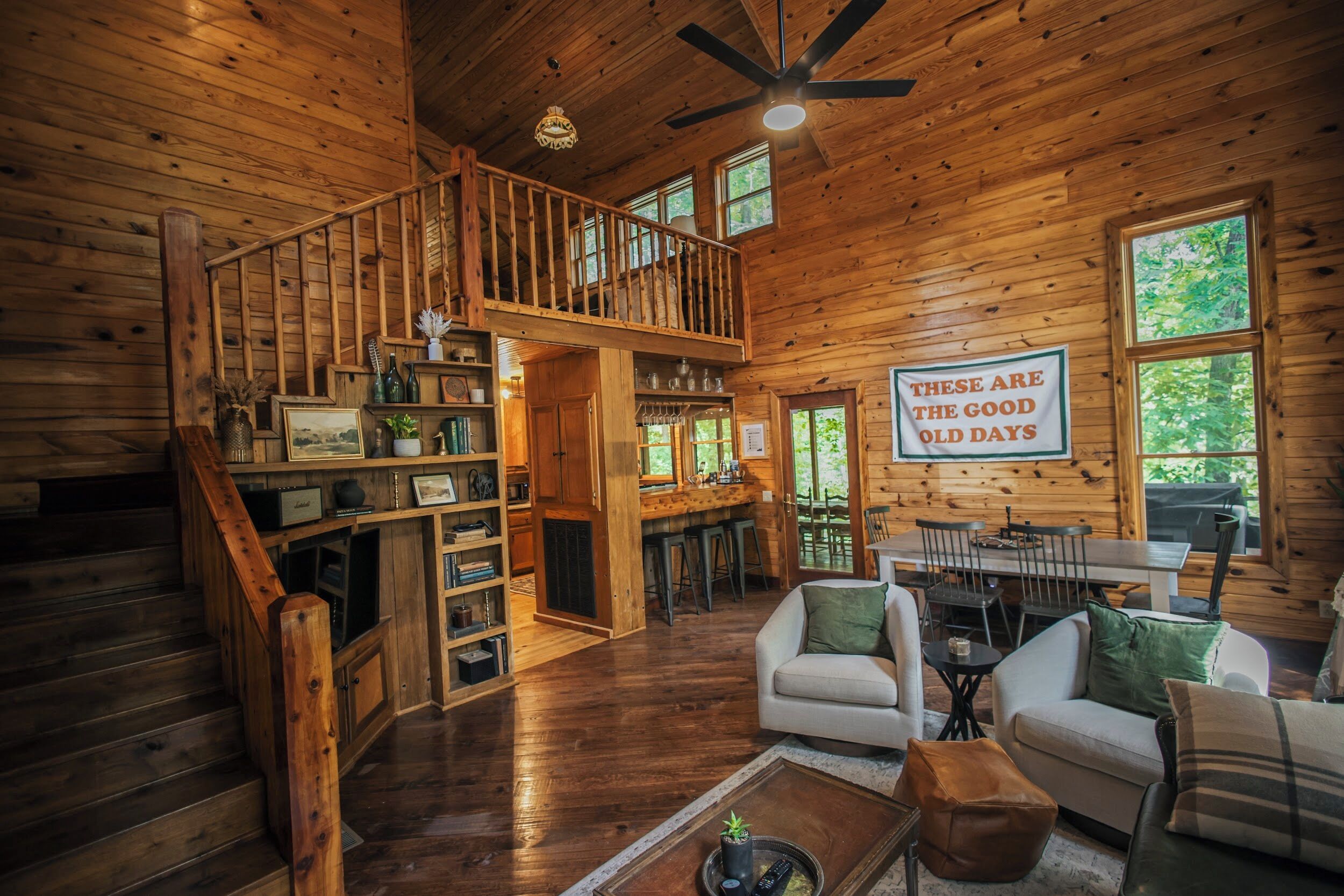 A wooden cabin interior with a staircase, a dining area, and a sign that reads 'These are the good old days.'