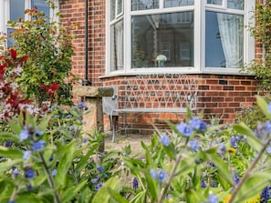 A brick building with a white window, a metal bench, and a garden with various plants. - 2 Bed in Filey (oc-49508) (Filey)