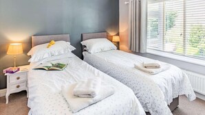 Une chambre avec deux lits, une table de chevet avec une lampe, une fenêtre avec des stores et une vue sur la verdure à l’extérieur.