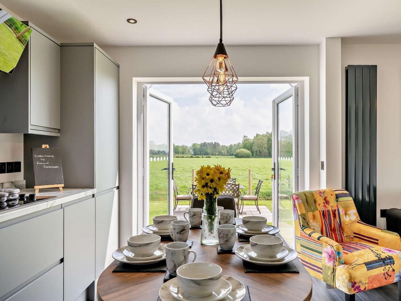 A modern kitchen with a dining area, a view of a green field, and a colorful chair.