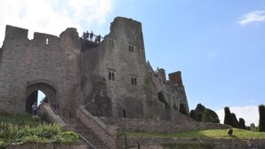 A historic stone castle with a large archway entrance, a staircase leading up to the main building, and a clear sky above.