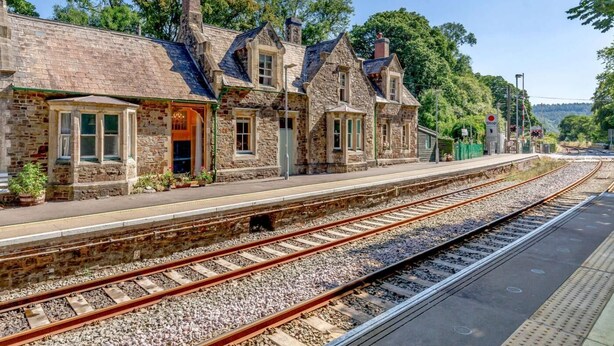 Edifici de pedra d'estil arquitectònic tradicional, una plataforma amb vies de ferrocarril i un senyal de 'Stop' que indica un pas de tren.