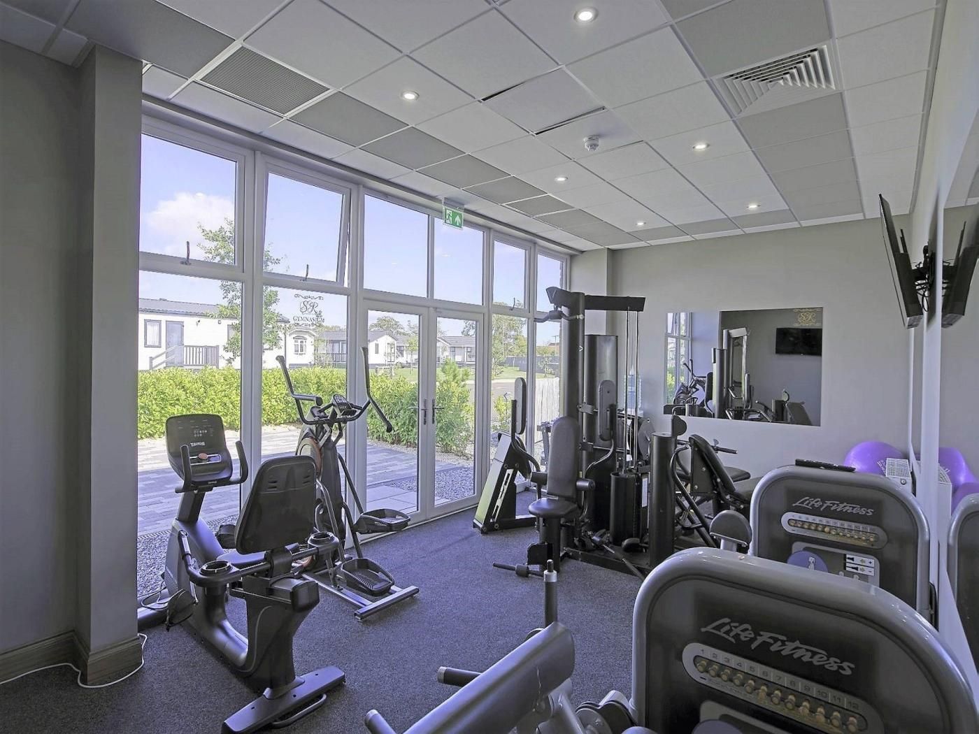 Ett gym med konditionsmaskiner, styrketräningsutrustning och stora fönster med utsikt över omgivningen.